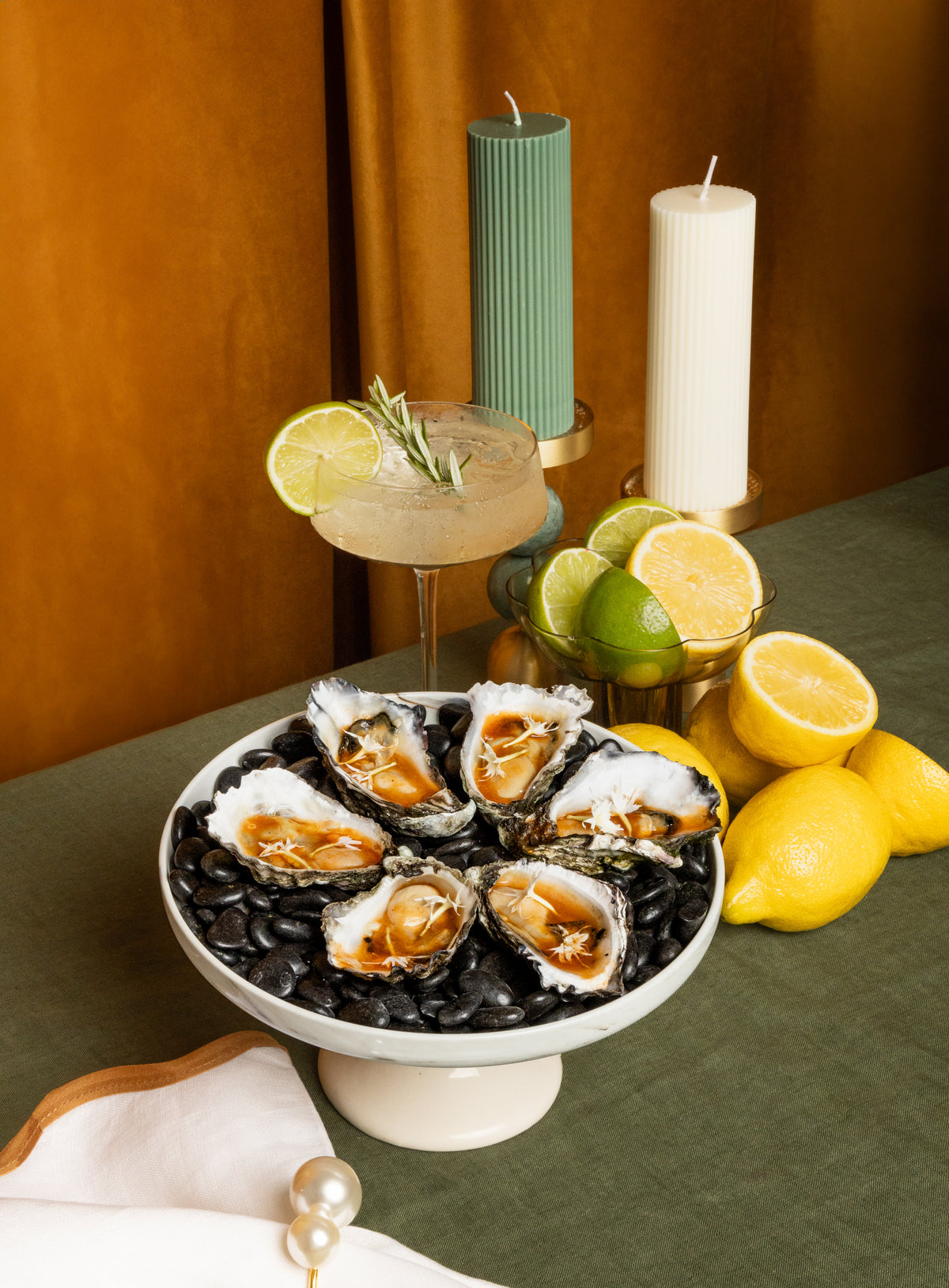 A styled food scene showing a platter of raw oysters on black beans, with a margarita glass, candles, limes, and lemons arranged on a table against a warm brown backdrop.