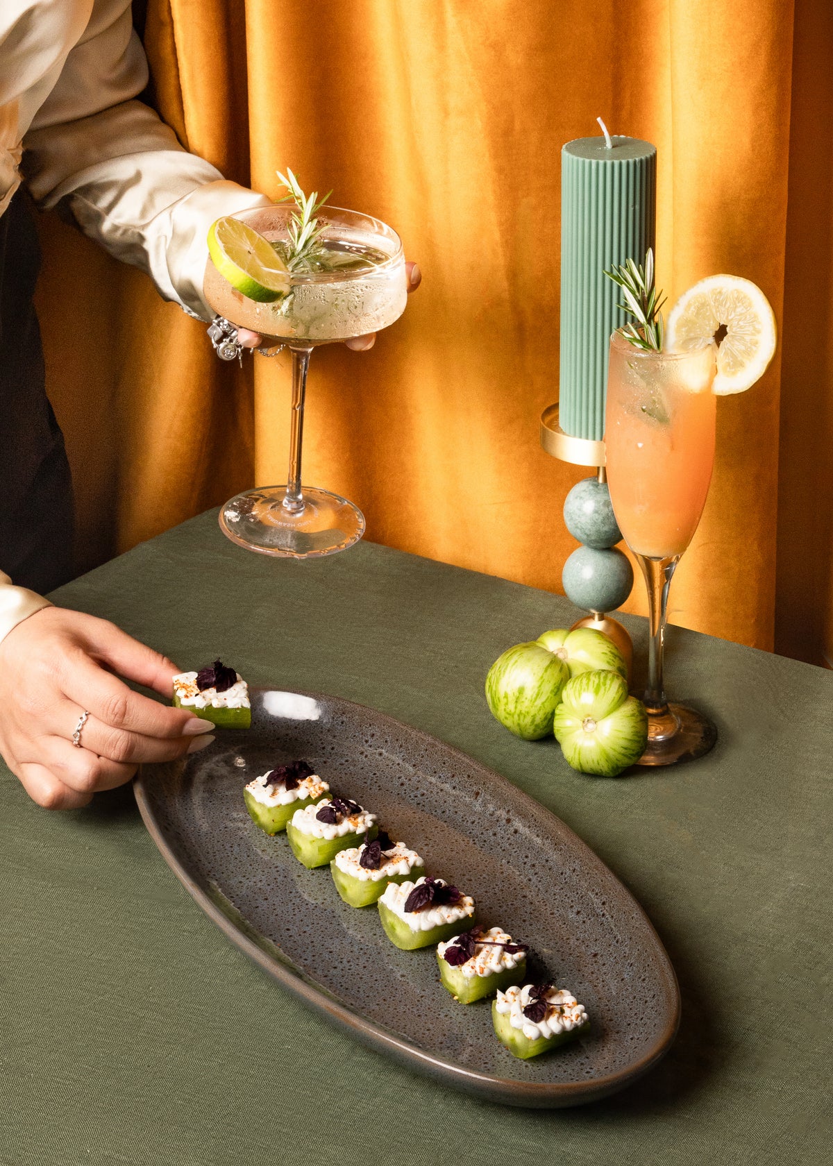 A close-up of a plated appetizer arrangement on a dark oval platter, with a person's hands holding a cocktail glass in the background against a warm orange curtain. The platter holds several small green appetizers topped with white cream and dark fruit preserves, with additional garnishes and green apples on the table.