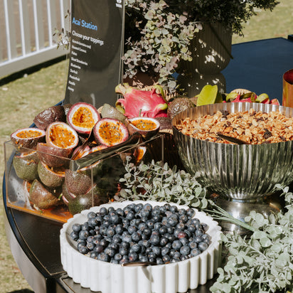 Assorted fruits and a bowl of berries on a table with decorative elements. Acai Bra. Acai Cart, Acai Toppings.