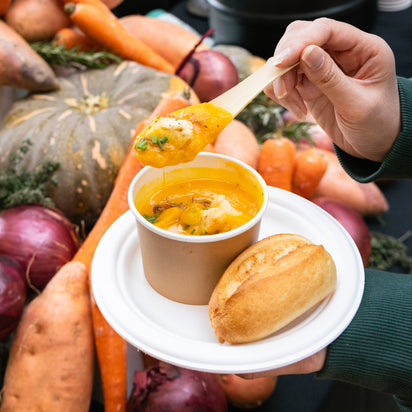 Person holding a plate with pumpkin soup and bread, surrounded by vegetables