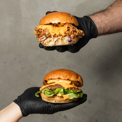 Two burgers held by hands in black gloves against a grey background