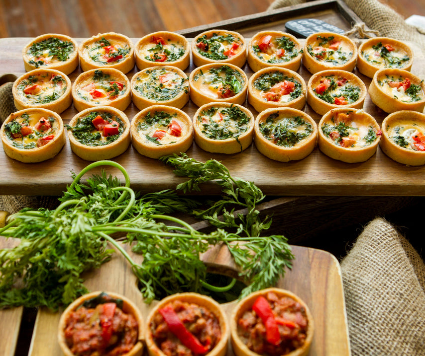 A wooden serving board holds rows of small baked tartlets filled with herbs, cheese, and red peppers, arranged on a rustic table with burlap and additional garnish in the foreground.