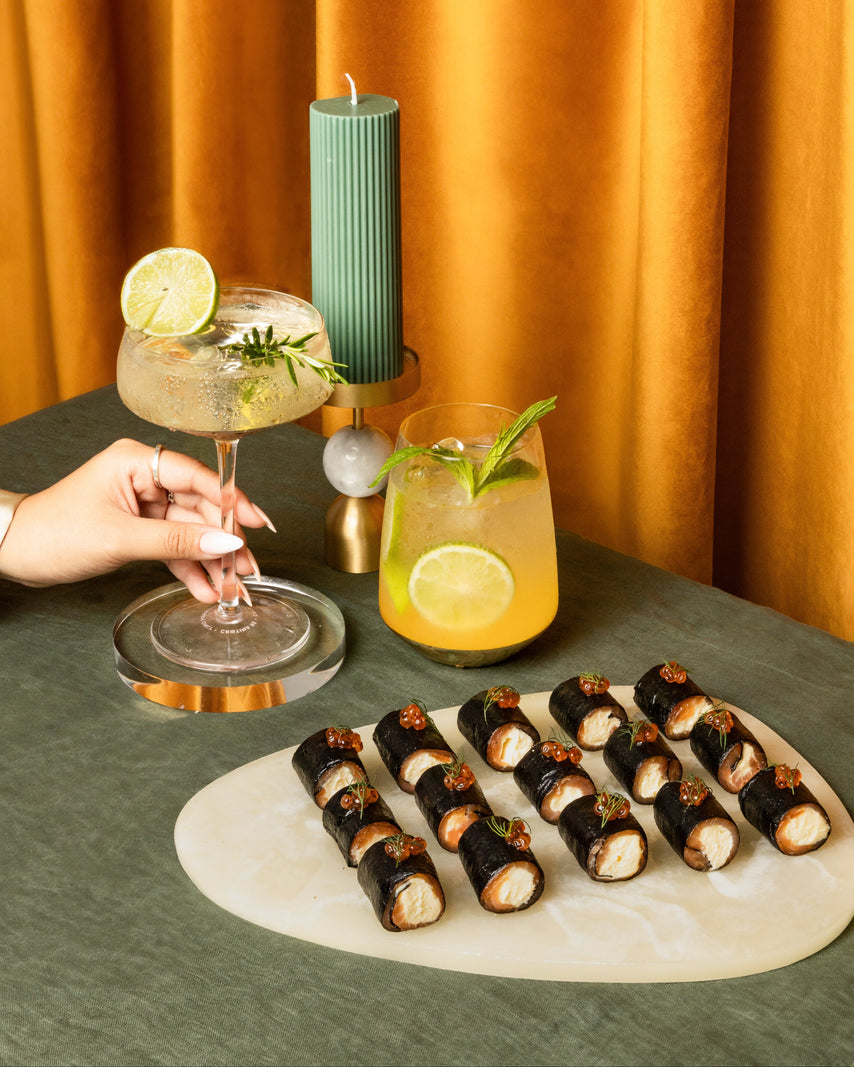 A styled tabletop scene with a hand holding a coupe glass filled with a pale yellow cocktail and a lime wheel, next to a glass of orange-yellow drink with mint. In front is a platter of sushi rolls on a white oval board, set against a dark green tablecloth and a warm orange curtain backdrop.