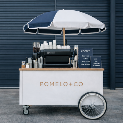 Coffee cart with 'Pomelo+Co' branding under an umbrella against a dark wall.