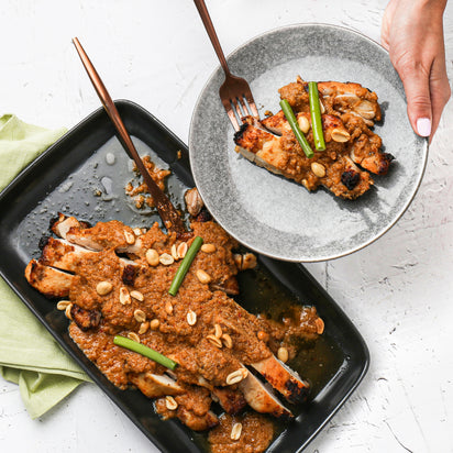 Baked chicken with peanut sauce on a black tray and gray plate, with a hand holding the plate.