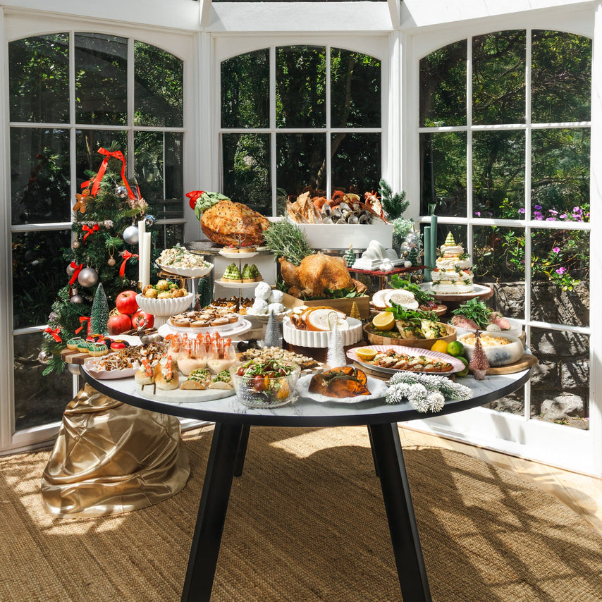 A round black dining table laden with an abundant holiday feast, set inside a bright conservatory with large windows and a glass pyramid roof. A decorated Christmas tree stands to the left.