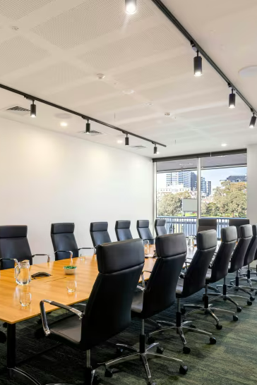 A modern conference room with a long rectangular wooden table surrounded by black office chairs, large windows overlooking a balcony and city view, and track lighting on the ceiling.