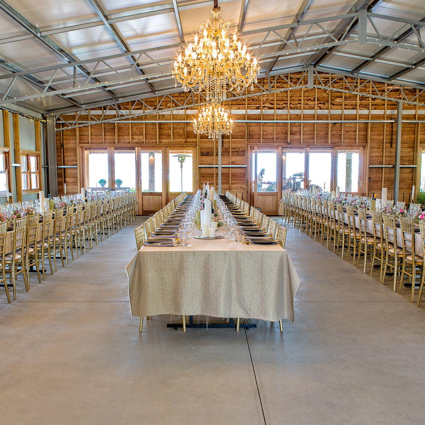 A large indoor event space with a high metal and wood ceiling, multiple hanging chandeliers, and long dining tables covered in cream tablecloths and surrounded by chairs, set up for a birthday party.