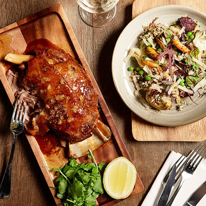 Roasted leg of lamb on a wooden platter with a side salad on a wooden table.