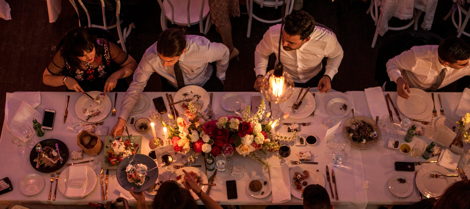 Overhead view of a formal dinner table setting with multiple guests seated around a long table decorated with floral centerpieces and lit candles in a dimly lit event space.