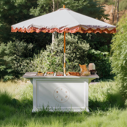 Outdoor setup with a white table under a large umbrella, surrounded by greenery. Grazing Cart.