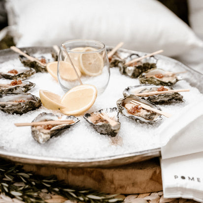 Oysters on a bed of ice with lemon wedges and a glass of wine, set on a white surface.