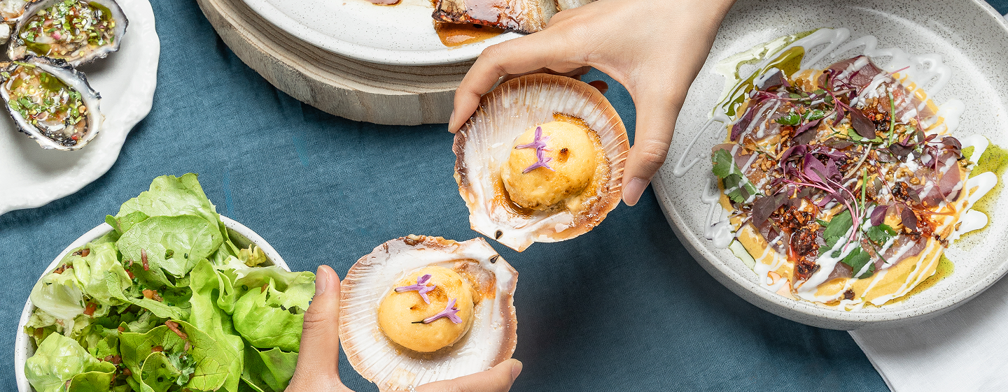 Two people cheers there scallops over a blue table filled with dishes.