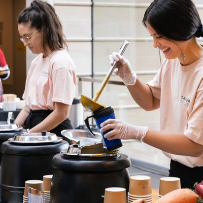 Two women in pink shirts and gloves preparing food in a kitchen.