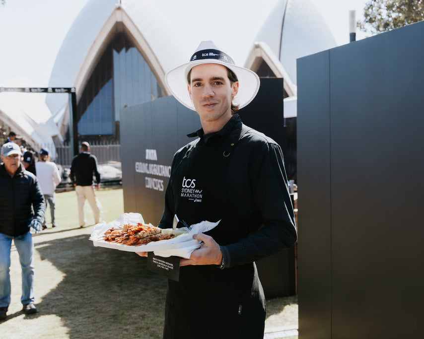 A man in a black chef jacket and white hat holds a tray of cooked chicken wings in front of the Sydney Opera House, with a large black screen behind him displaying 'NSW NATIONAL CHICKEN CONTEST'.