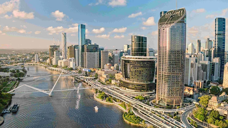 Aerial view of a Brisbane city skyline with a river running through the foreground, bridges crossing the water, and several tall skyscrapers under a blue sky with scattered clouds.